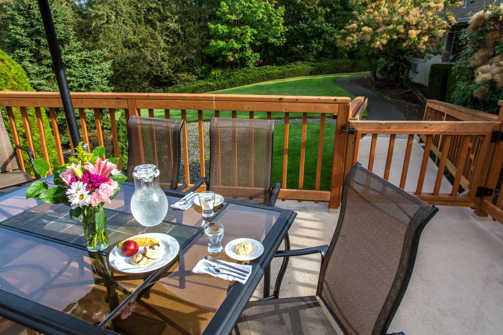 A patio deck setup with a dining table and chairs, featuring a meal and flowers overlooking a garden view.