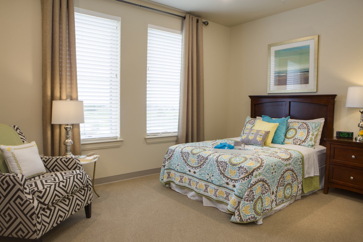A cozy bedroom with a neatly made bed, decorated with patterned bedding, beside two windows with curtains and a lamp on the side table.