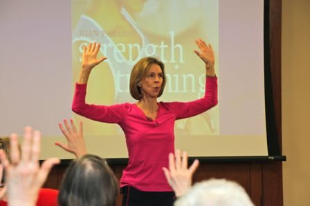 A woman in a pink shirt raises her hands during a presentation in front of an audience.