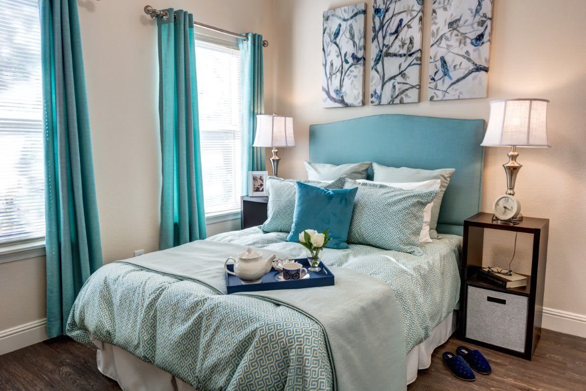 A cozy bedroom featuring blue-toned decor, elegant artwork, and neatly arranged bedding.