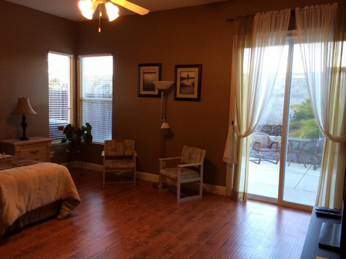 A cozy bedroom with wooden flooring, featuring two chairs, a bed, and large windows with curtains allowing natural light to fill the room.
