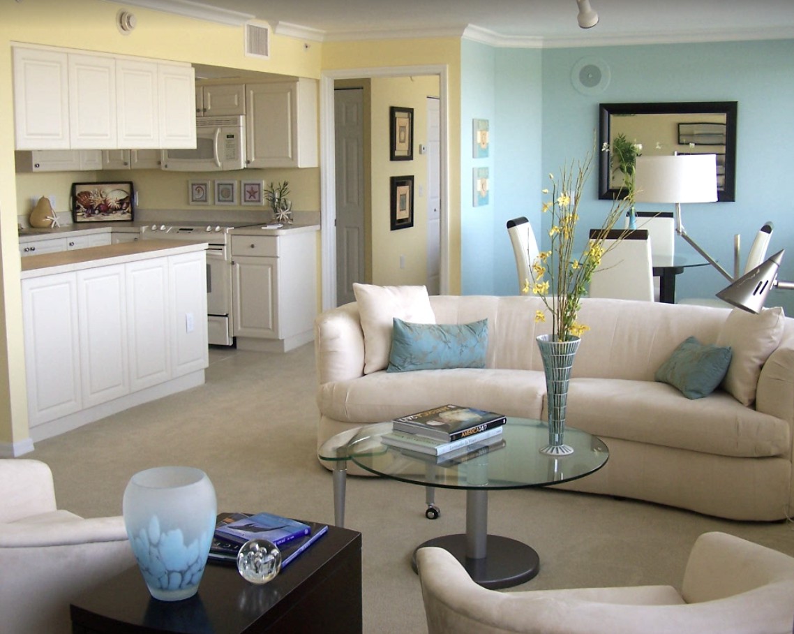 A modern living room with a beige sofa set, glass coffee table, and an open kitchen in the background.