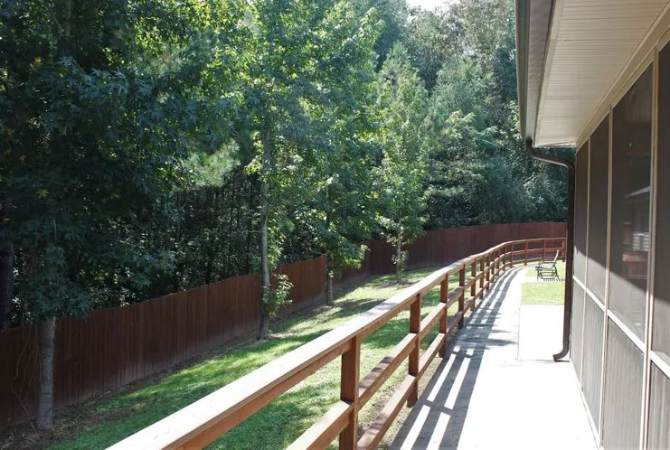 A sunny backyard with a wooden deck and railing overlooking trees and a fence.