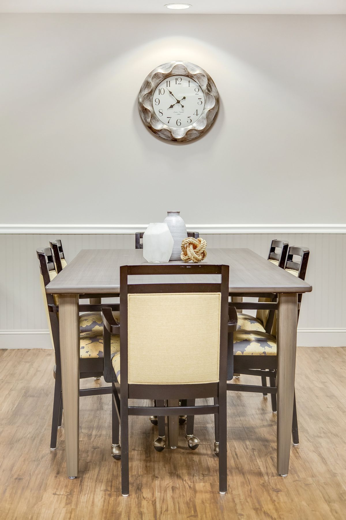 A dining room with a wooden table, matching chairs, and decorative elements including a wall clock and vases.