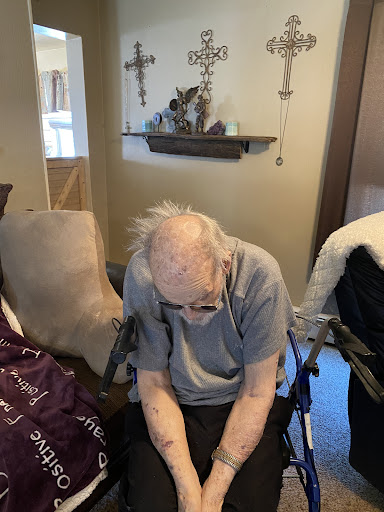 An elderly person sitting in a wheelchair, appearing thoughtful or reflective in a cozy indoor setting.