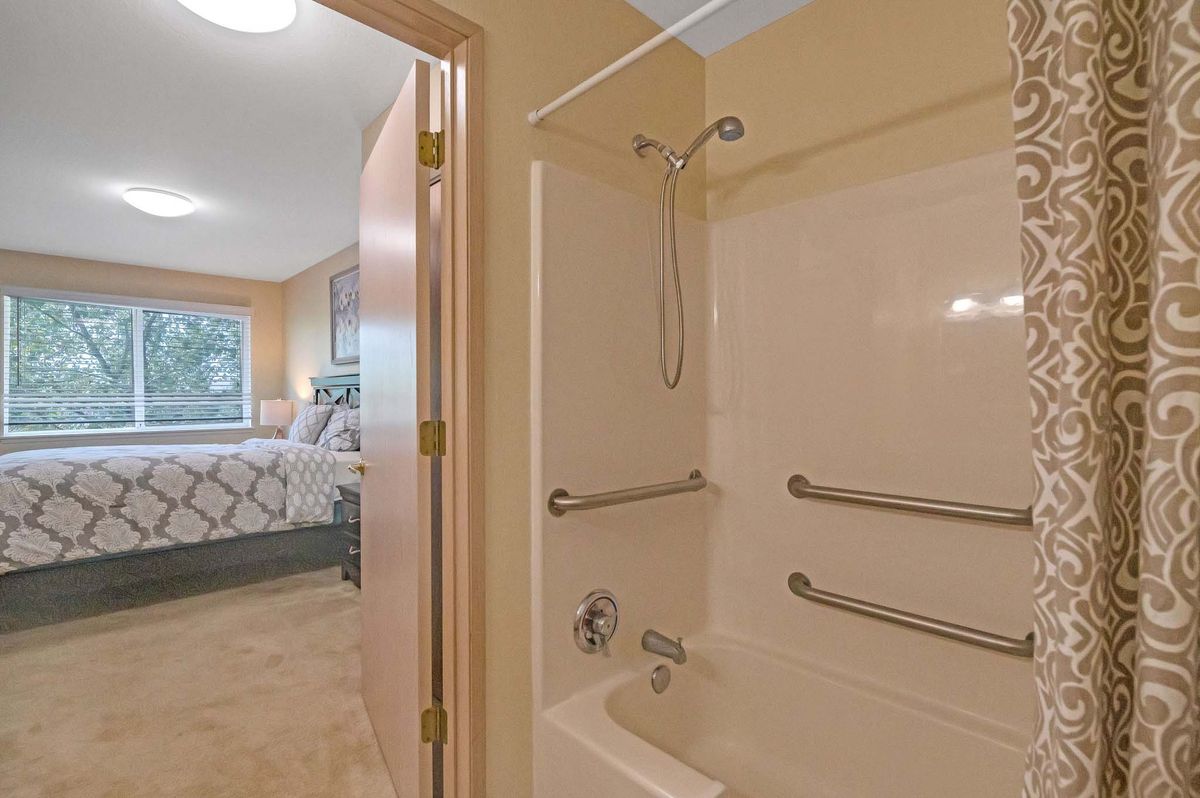 A view from a bathroom with a bathtub and grab bars, leading into an adjacent bedroom.