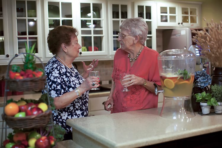 Two elderly women having a conversation in the kitchen while holding drinks.