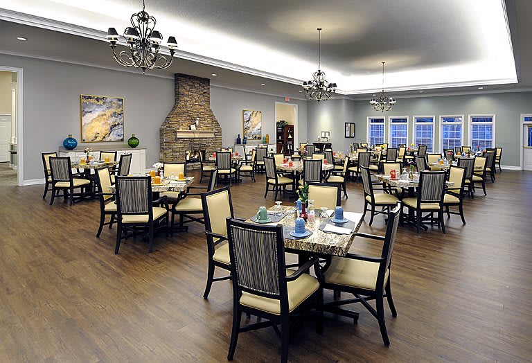 An elegant dining room with neatly arranged tables and chairs, featuring chandeliers and a fireplace.