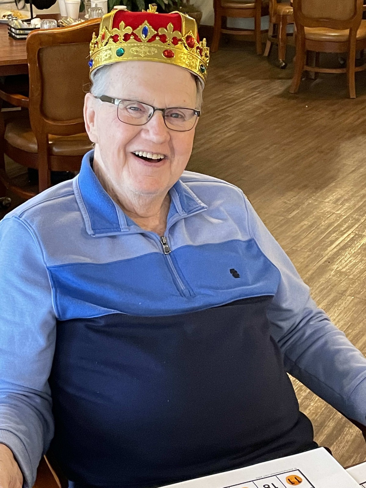 An elderly man with glasses is smiling while seated indoors, wearing a small crown.