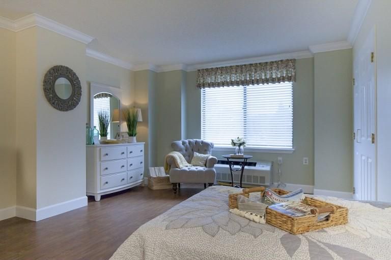 A cozy bedroom with a serene color palette and elegant furnishings, including an armchair, dresser, and window with soft natural lighting.