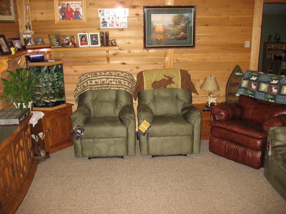 A cozy living room with wooden walls, featuring two green recliners, a leather armchair, a fish tank, and various decorative elements.