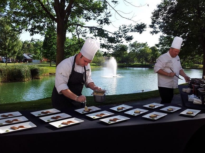 Chefs meticulously plating dishes at an outdoor event by a scenic lake and fountain.