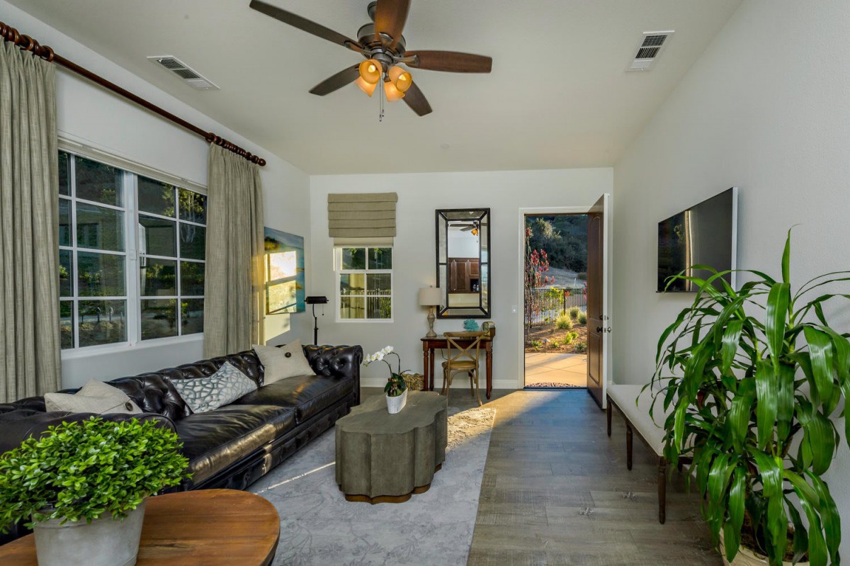 A cozy living room featuring a leather sofa, coffee table, ceiling fan, and large windows letting in natural light.