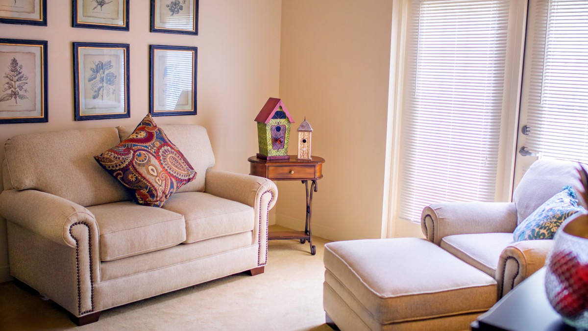 A cozy living room with a sofa, patterned cushions, and an ottoman beside a window. A small table holds colorful birdhouses as decor.