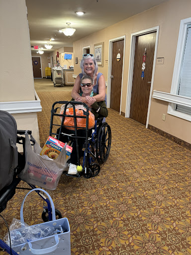A caregiver assists an elderly individual in a wheelchair down a hallway.
