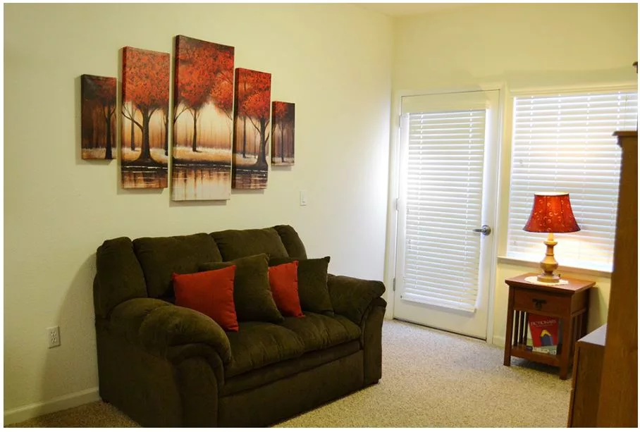 A cozy living room featuring a dark green sofa with red pillows, adorned with multi-panel wall art depicting trees. Natural light pours in through the window beside a small wooden table with a red lamp.