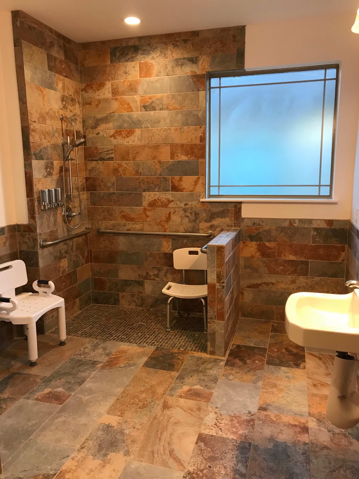 An accessible bathroom with a stone tile design, featuring a shower area, seat, and sink.