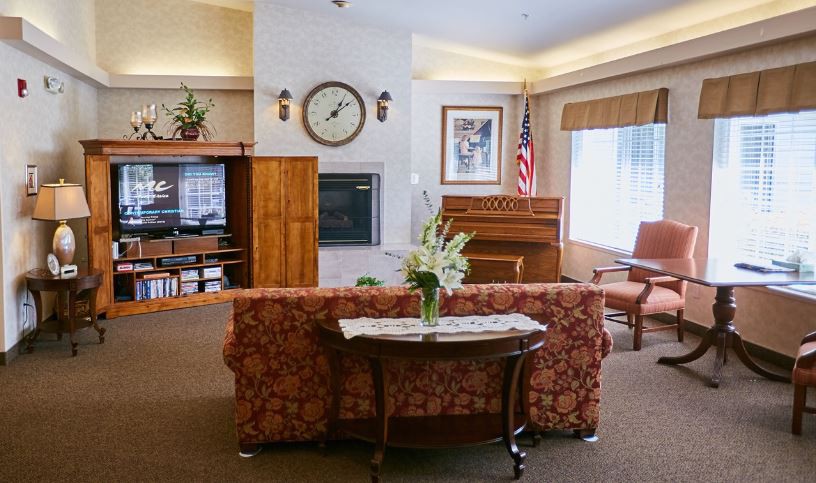 A cozy living room with a piano and floral decorations, featuring a large clock and comfortable seating.