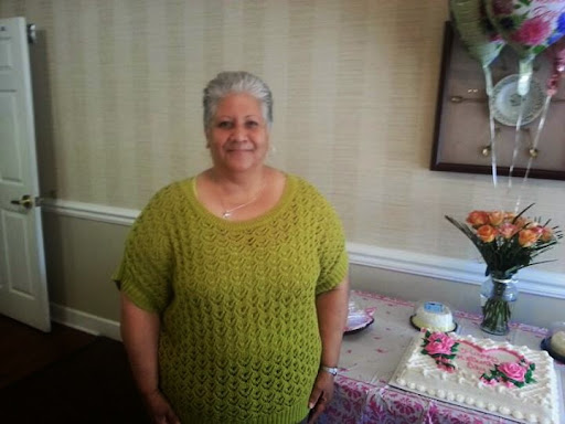 A joyful woman in a green sweater stands beside a beautifully decorated birthday cake.