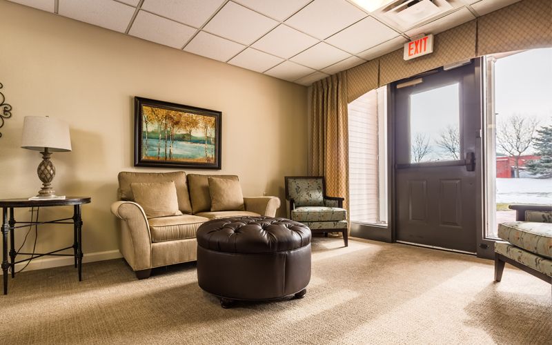 A cozy waiting room featuring a sofa, armchair, and ottoman, with a painting on the wall and a carpeted floor.