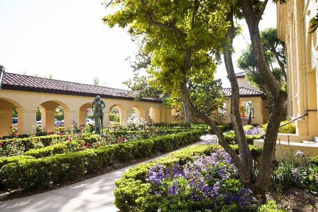 A tranquil garden with a pathway lined by blooming flowers and shaded by trees, alongside an arched wall.