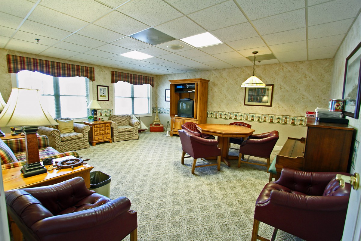 A cozy living room with comfortable seating and a central table, featuring a patterned carpet and ample natural light from the windows.