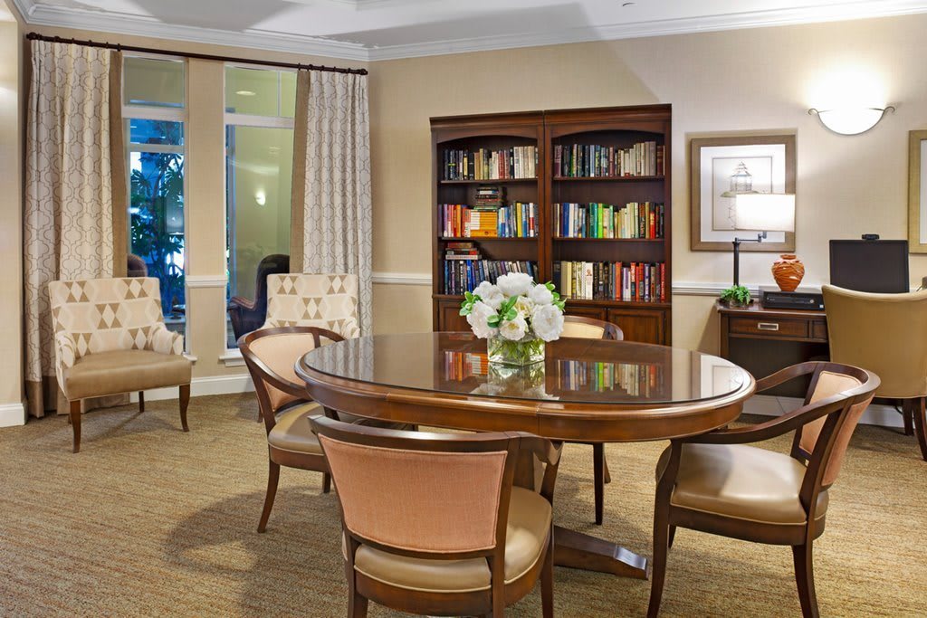 A cozy reading area featuring a polished wooden table surrounded by chairs, with a bookshelf full of books in the background and a vase of flowers on the table.