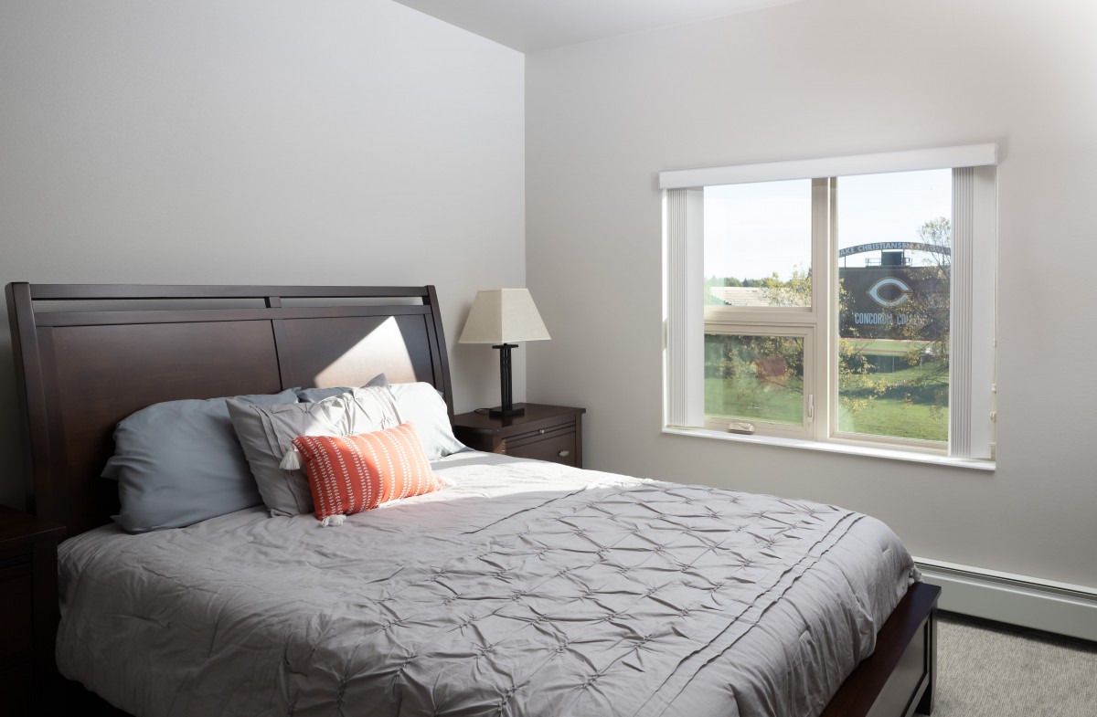 A cozy bedroom with a neatly made bed, featuring colorful pillows and a bedside lamp next to a window.