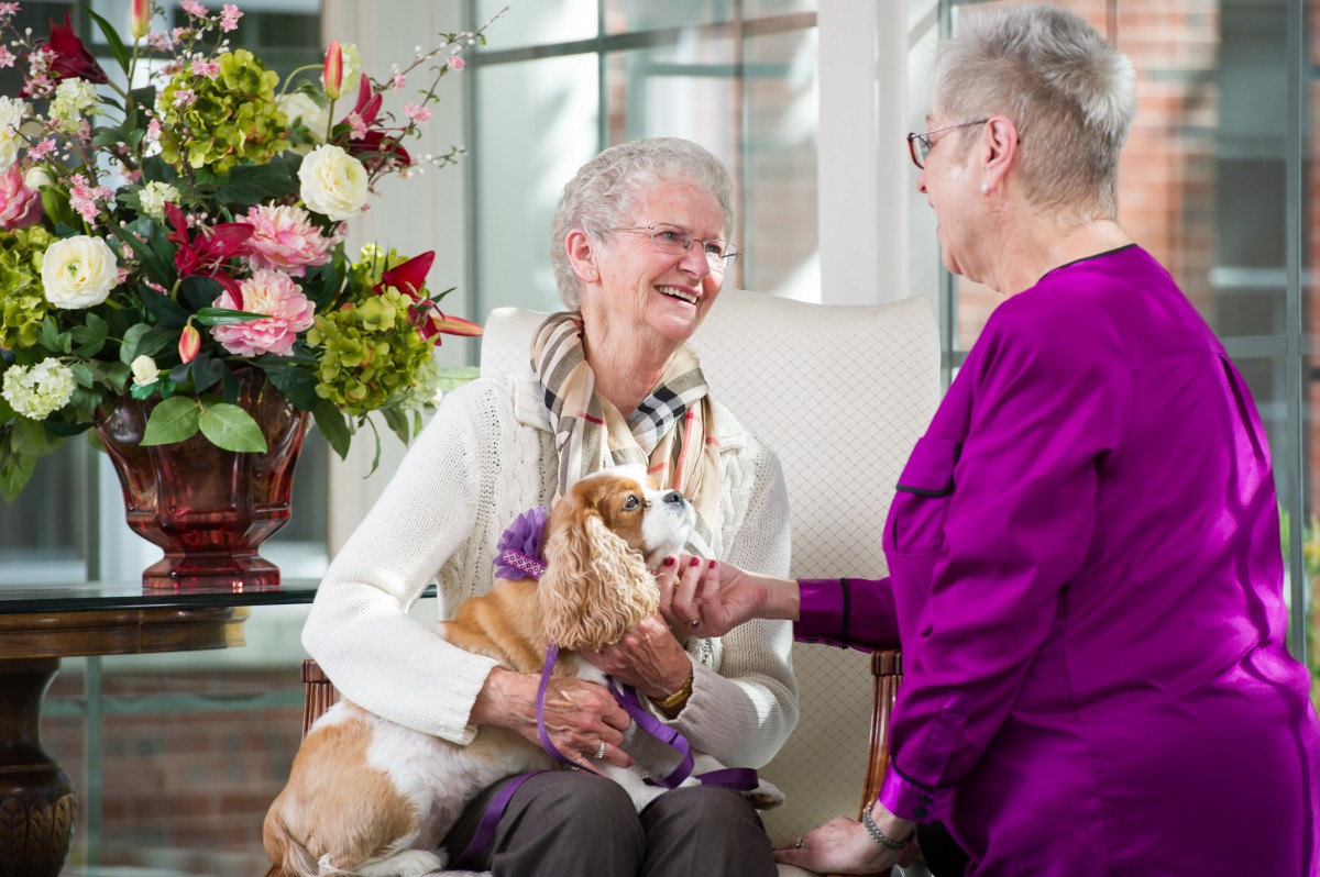 Two elderly women smiling and conversing while one holds a dog, with flowers in the background.