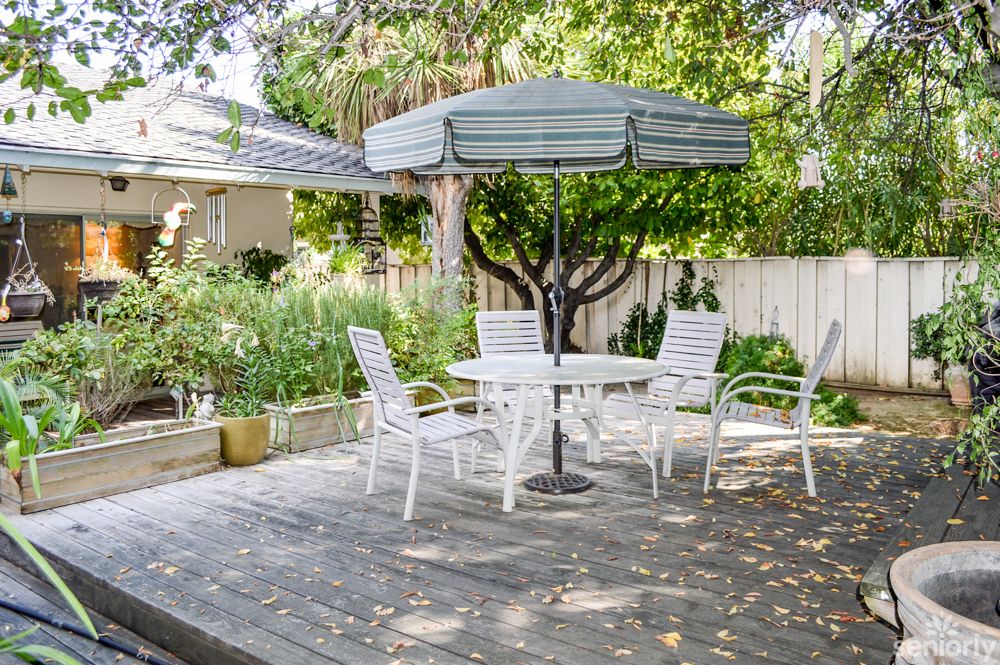 A sunny backyard patio featuring a table with an umbrella and several chairs, surrounded by lush greenery.