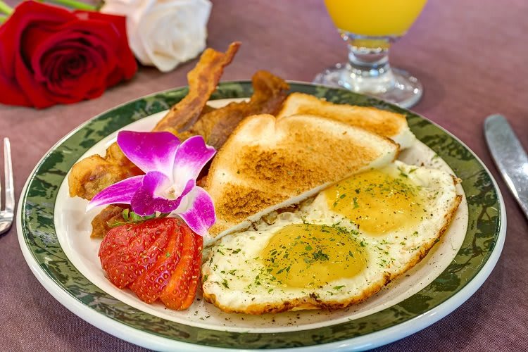 A delicious breakfast plate featuring sunny-side-up eggs, crispy bacon, toast, a sliced strawberry, and a decorative orchid flower, accompanied by a glass of orange juice.