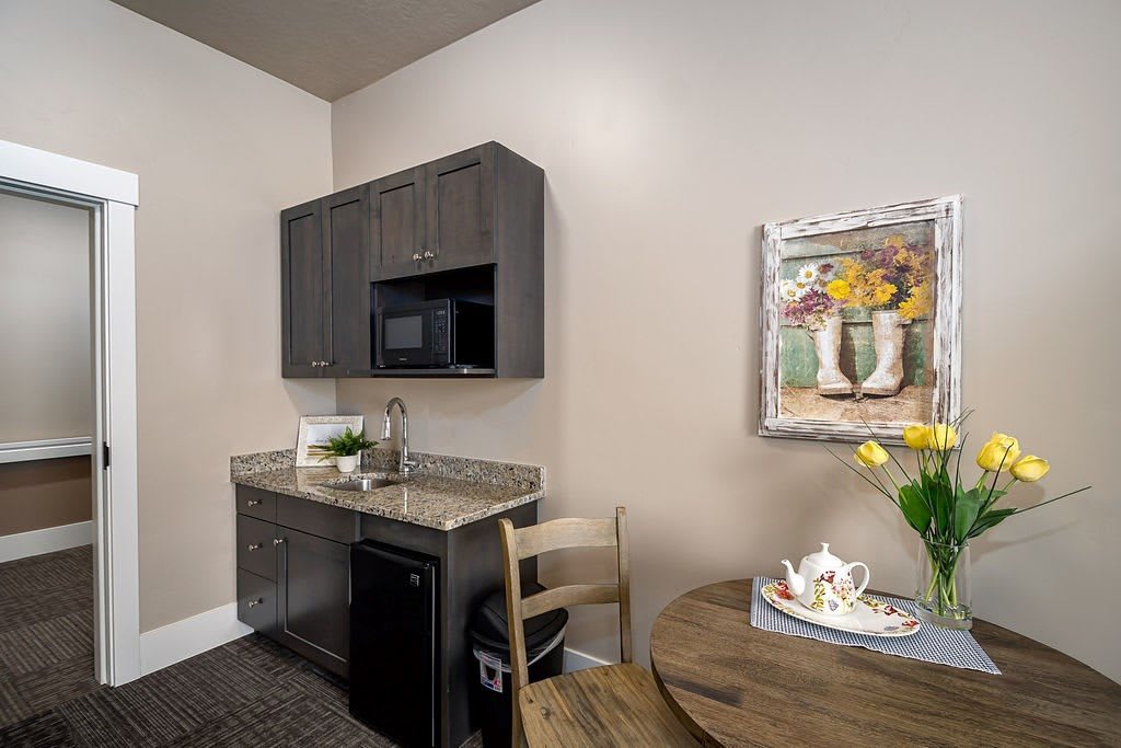 A cozy kitchenette with dark cabinets, a small sink, and a microwave, featuring a round table with seating and decorative elements.