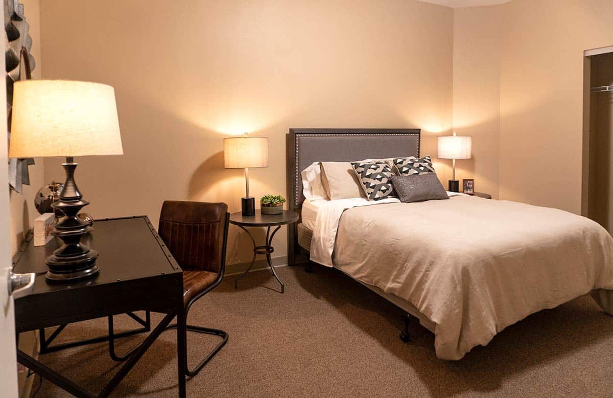 A cozy and minimalist bedroom with a comfortable bed, soft lighting from lamps, and a desk with a chair.