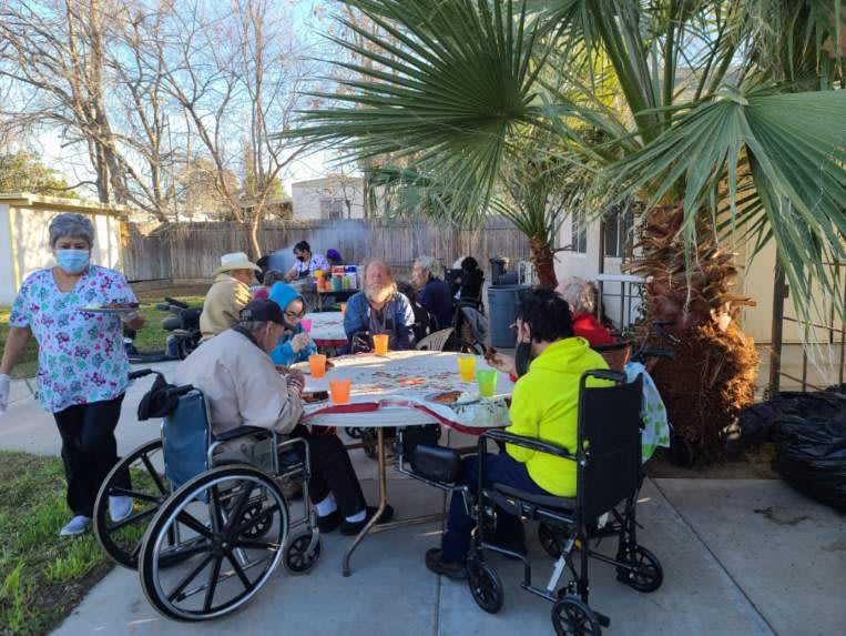 A community gathering with people of different ages enjoying an outdoor social event under a palm tree.