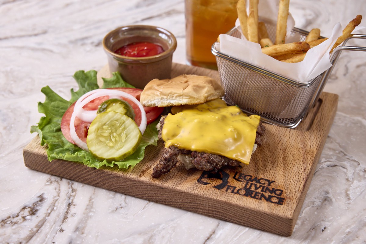 A cheeseburger served with lettuce, tomato, onion, and pickles on a wooden board, accompanied by a basket of French fries and a small bowl of ketchup.