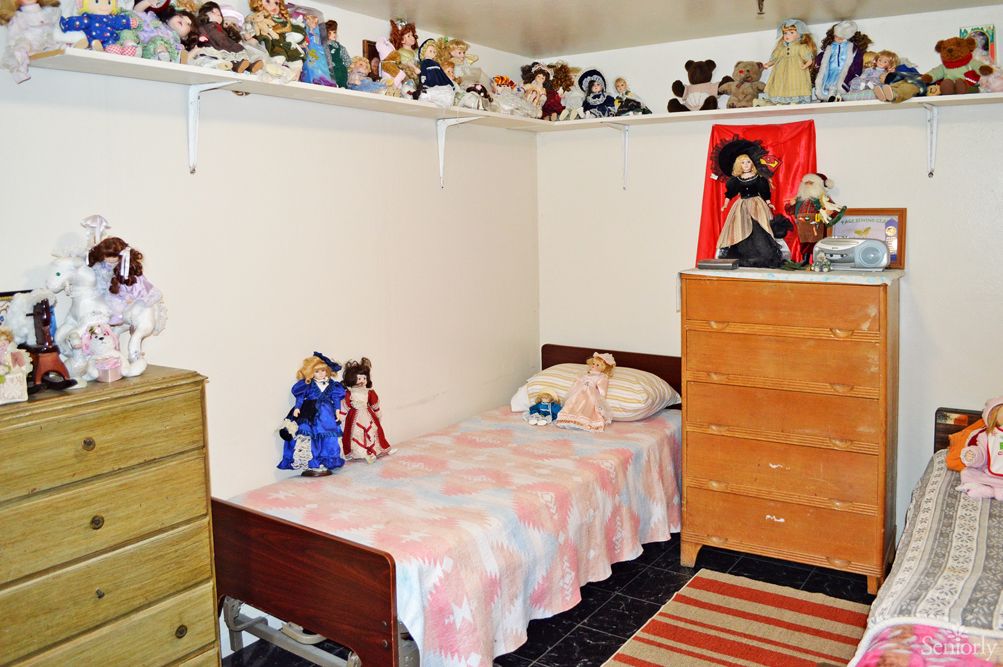 A cozy bedroom featuring a single bed with dolls and a dresser, adorned with numerous toys on shelves.