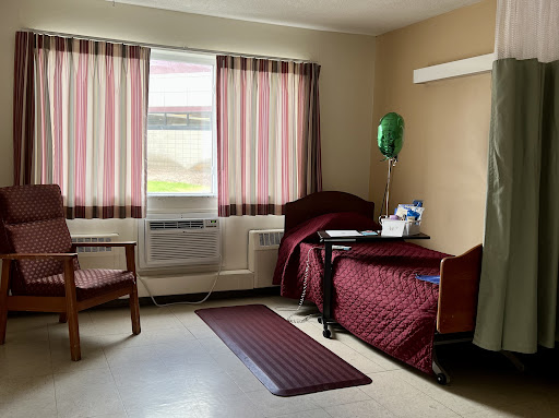 A hospital room featuring a bed with maroon bedding, two chairs, and striped curtains.