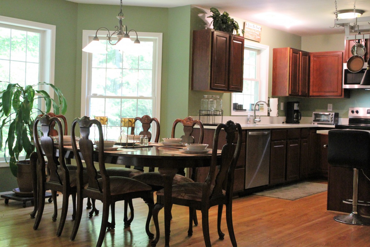 A cozy dining area with a wooden table and chairs, adjacent to a kitchen with dark cabinets and a large window.