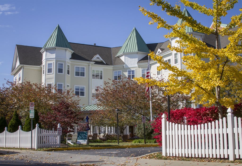 A picturesque house with green rooftops surrounded by autumn trees and a white picket fence.