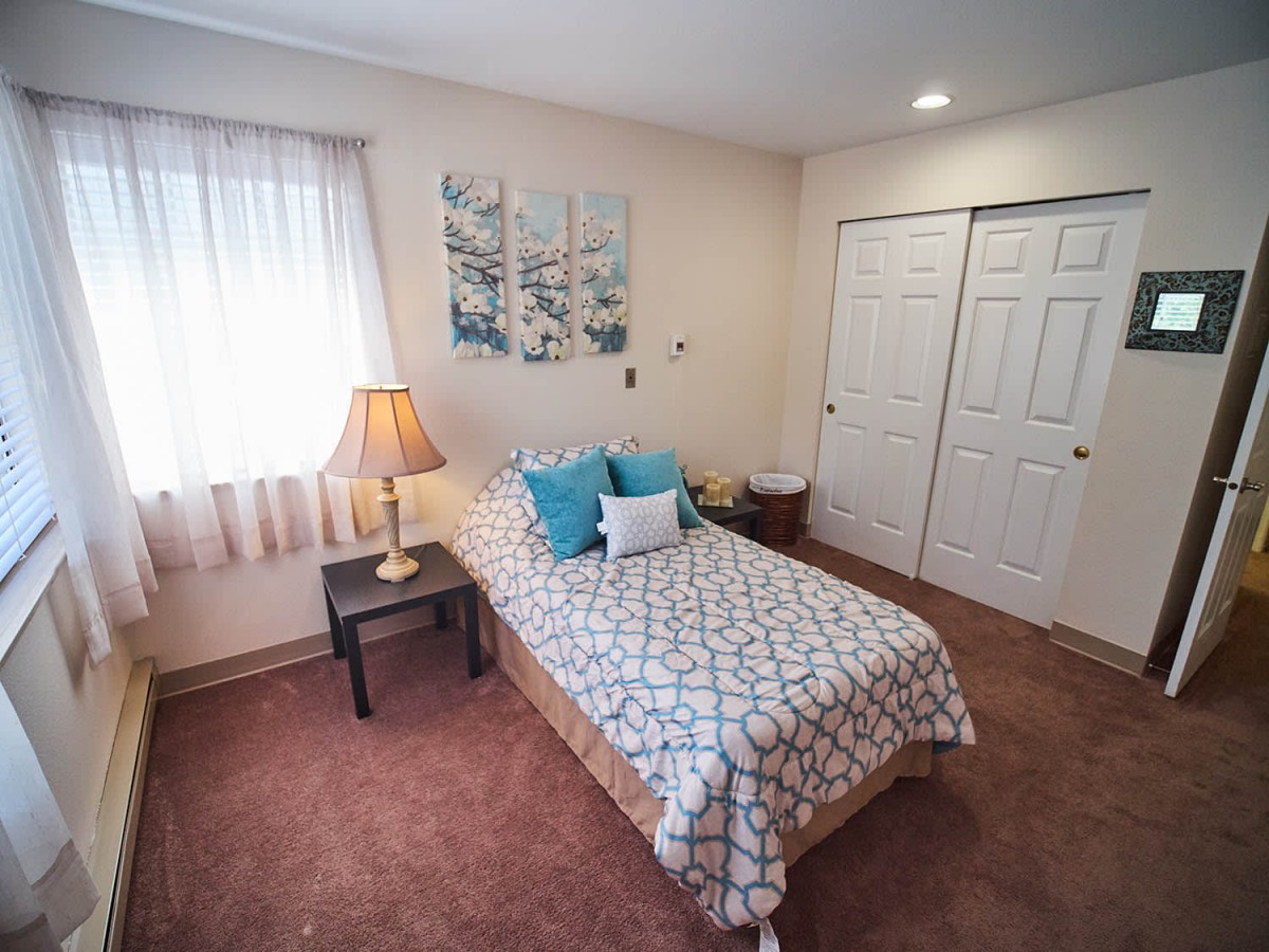 A cozy bedroom with a single bed, decorated with pillows and a bedside lamp, featuring a carpeted floor and a closet.