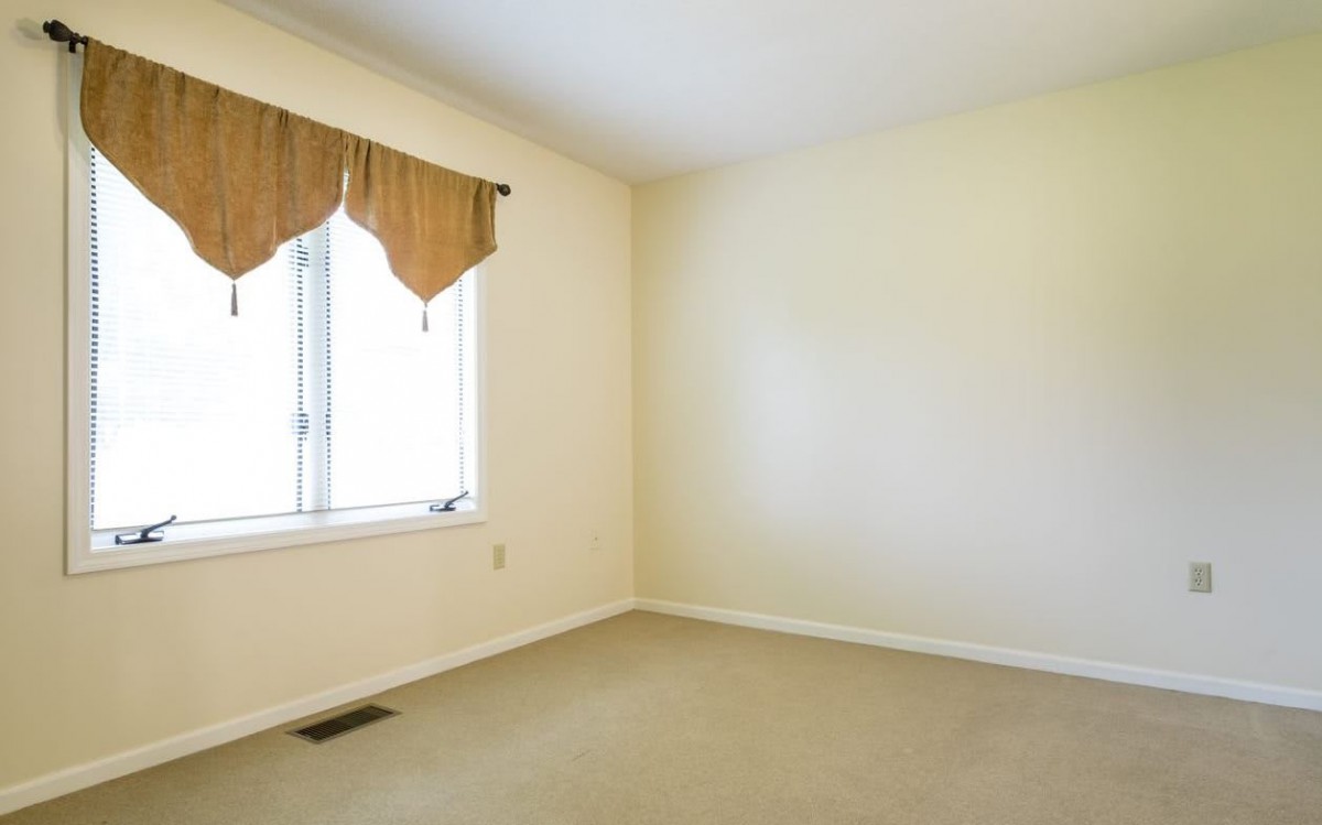 An empty room with beige walls, carpet, and a window with curtains.