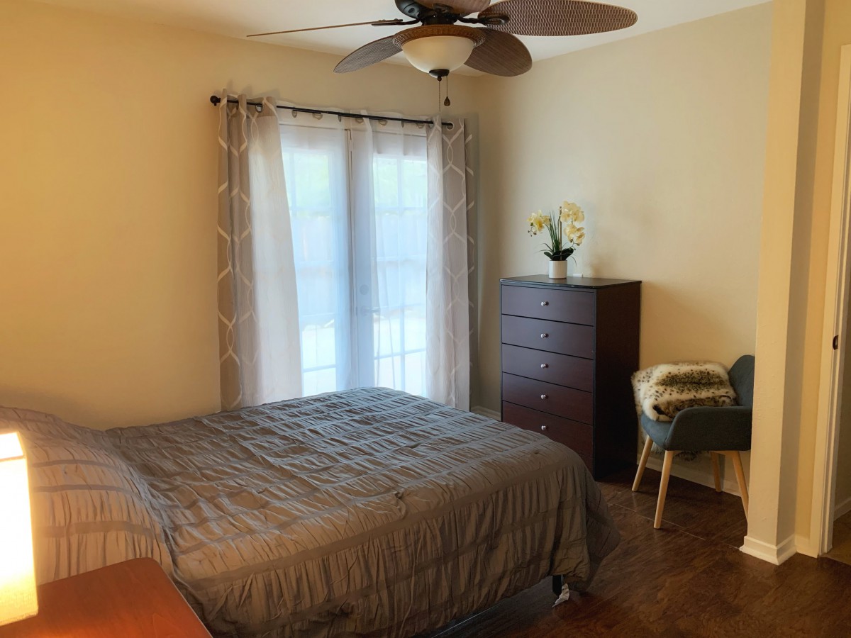 A cozy bedroom featuring a bed with a grey comforter, wooden dresser, chair, and ceiling fan, with light streaming through the window curtains.