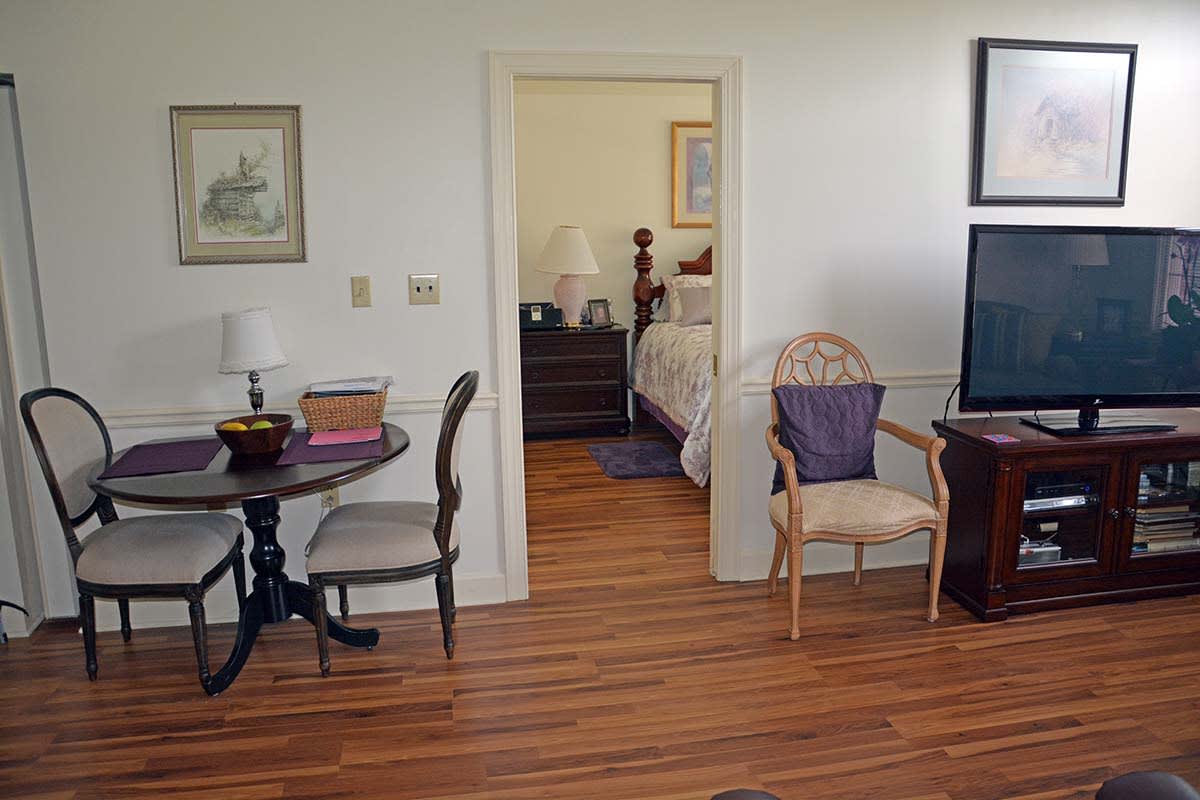 A view of a living room leading into a bedroom, featuring wood flooring and various furniture pieces.