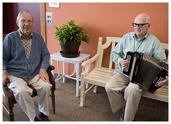 Two elderly men seated indoors, one playing an accordion.