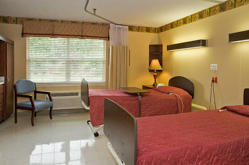 A cozy patient room featuring two beds, a lamp, and a large window for natural light.
