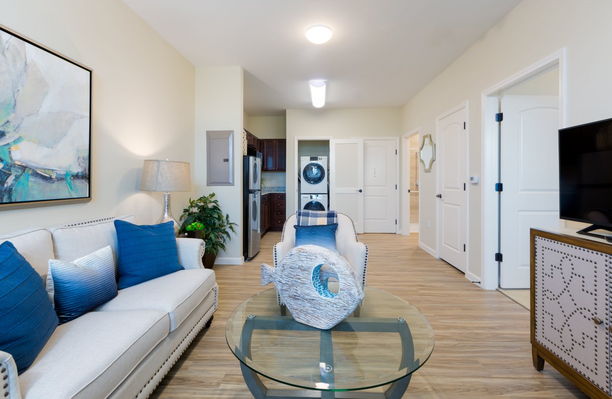 A modern living room with elegant decor, featuring a white sofa, glass table, and view into the kitchen.