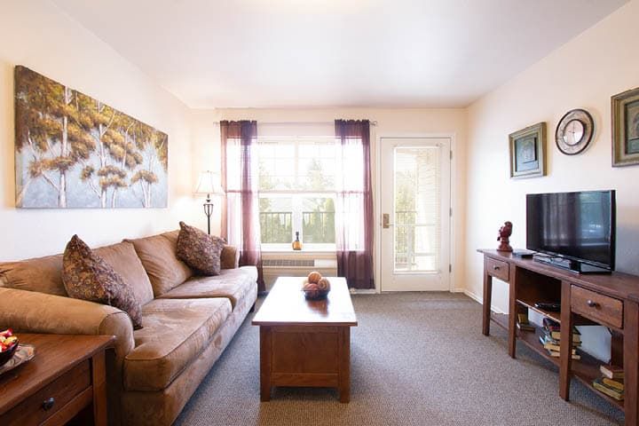A cozy living room with a sofa, coffee table, and television, featuring a large window with curtains and decorative elements.