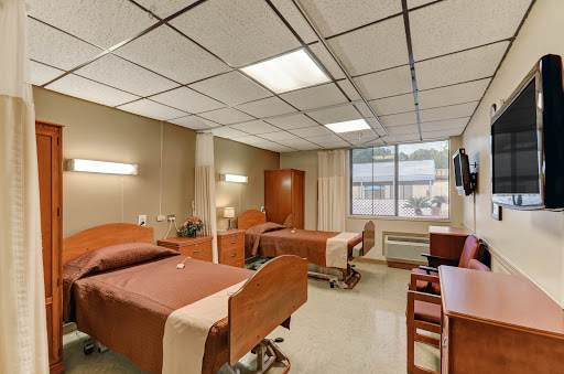 A view of a hospital patient room featuring two beds, essential furnishings, and natural light from a nearby window.