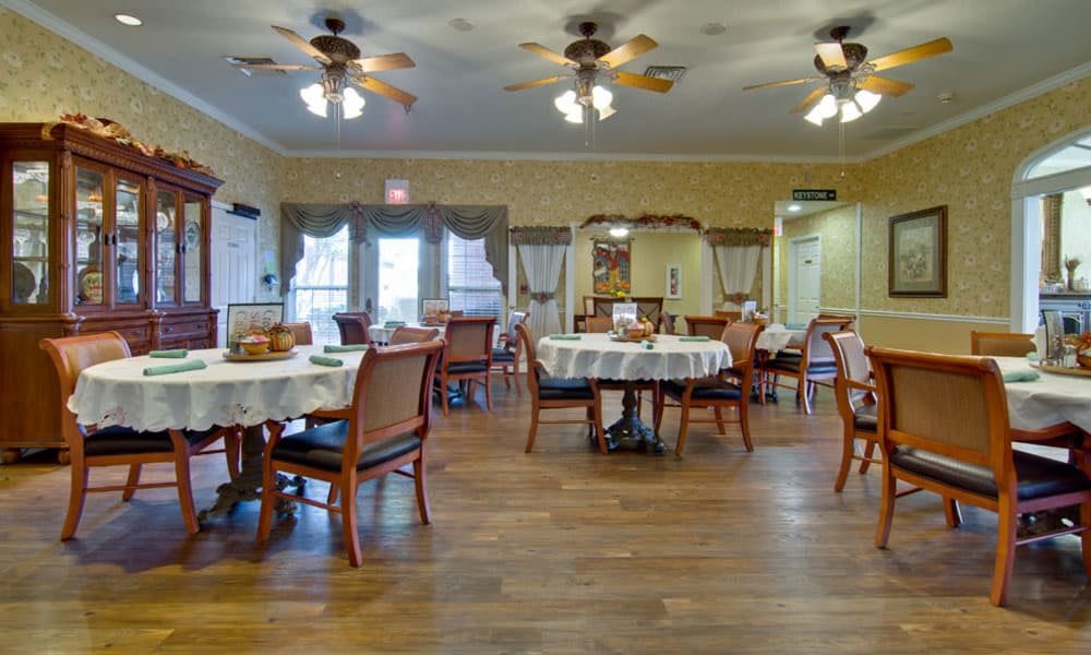 An elegant dining room with neatly arranged tables and chairs, featuring wooden flooring and ceiling fans.