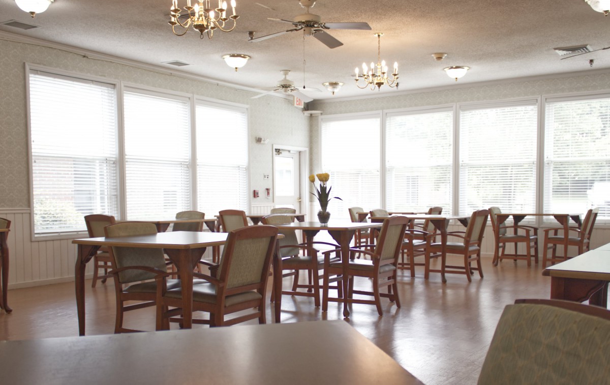 A bright and spacious dining room with multiple tables and chairs, illuminated by chandeliers and natural light from large windows.